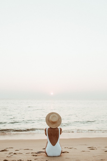 Jovem mulher bronzeada vestindo um lindo maiô branco com um chapéu de palha está sentada e relaxando em uma praia tropical com areia branca e está assistindo o pôr do sol e o mar