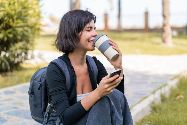 Jovem mulher búlgara bonita ao ar livre usando telefone celular e segurando um café para levar