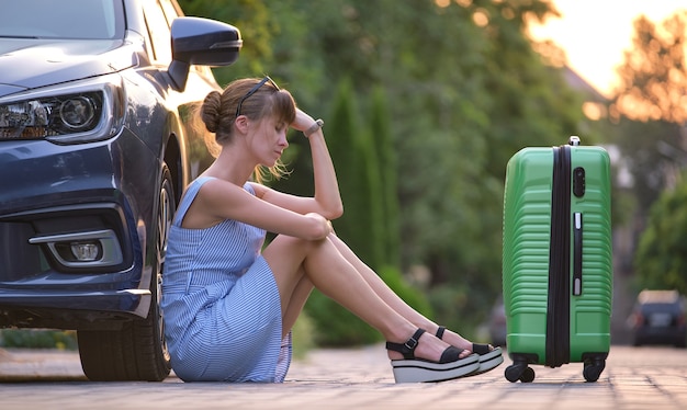 Jovem mulher cansada com mala sentada ao lado do carro, esperando por alguém. Conceito de viagens e férias.