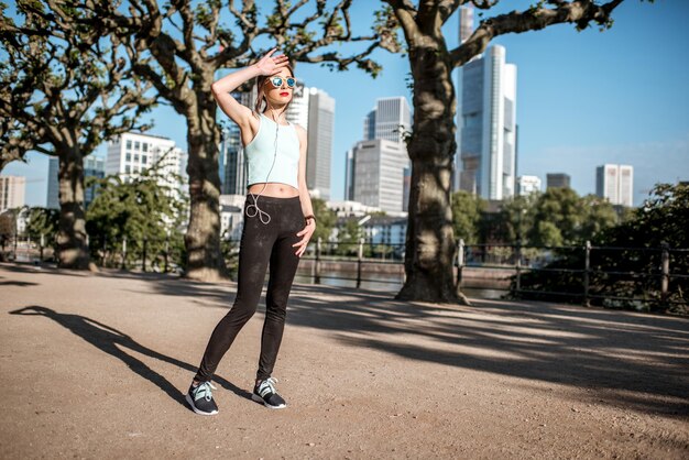 Jovem mulher cansada em roupas esportivas, fazendo um exercício matinal no parque com arranha-céus ao fundo na cidade de Frankfurt