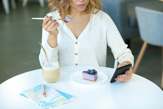 Jovem mulher casual rolando no smartphone enquanto está sentada à mesa no café e comendo um cheesecake de mirtilo