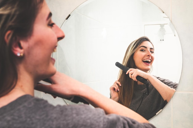 Jovem mulher caucasiana, alisando o cabelo com uma alisamento no espelho do banheiro.