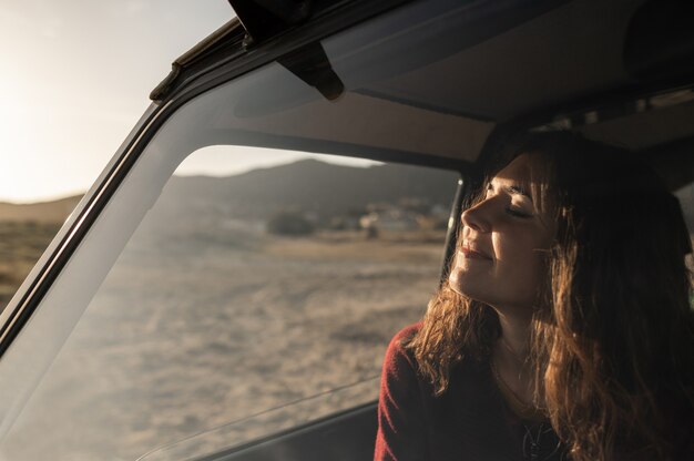 Jovem mulher caucasiana, aproveitando o sol com os olhos fechados, sentado dentro do carro, vista da janela. Conceito de viagem.