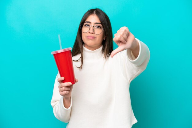 Jovem mulher caucasiana bebendo refrigerante isolado em fundo azul, mostrando o polegar para baixo com expressão negativa