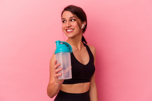 Jovem mulher caucasiana bebendo um shake de proteína isolado em fundo rosa olha de lado sorrindo alegre e agradável