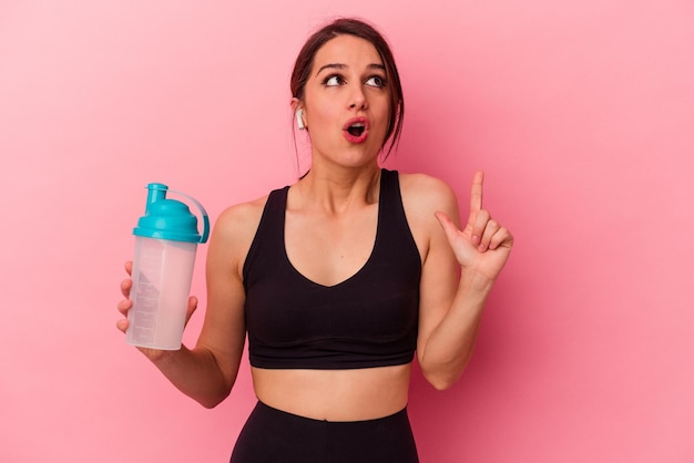 Jovem mulher caucasiana bebendo um shake de proteína isolado no fundo rosa apontando para cima com a boca aberta