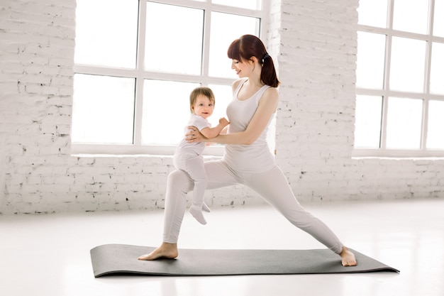 Jovem mulher caucasiana bonita fazendo exercícios de fitness no ginásio grande luz com sua filha criança juntos. Materialidade maternidade e maternidade