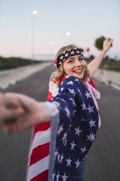 Jovem mulher caucasiana com cabelo loiro curto e uma bandeira dos EUA segurando uma mão enquanto caminhava na estrada