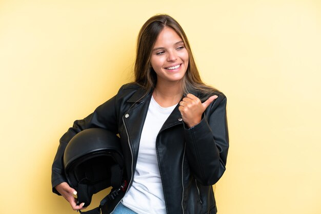 Jovem mulher caucasiana com capacete de moto isolado em fundo amarelo, apontando para o lado para apresentar um produto