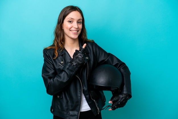 Jovem mulher caucasiana com capacete de moto isolado em fundo azul, apontando para o lado para apresentar um produto