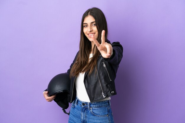 Jovem mulher caucasiana com capacete de motociclista isolada em fundo roxo sorrindo e mostrando sinal de vitória