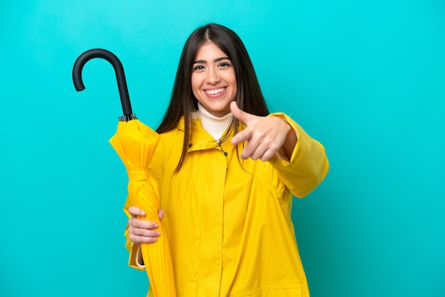 Jovem mulher caucasiana com casaco à prova de chuva e guarda-chuva isolado em fundo azul com polegares para cima porque algo bom aconteceu