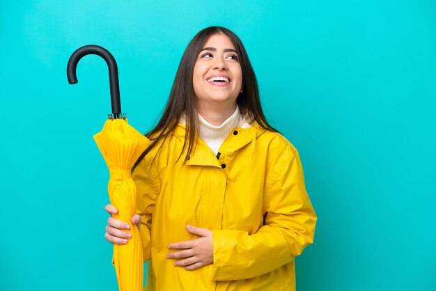 Jovem mulher caucasiana com casaco à prova de chuva e guarda-chuva isolado em fundo azul sorrindo muito