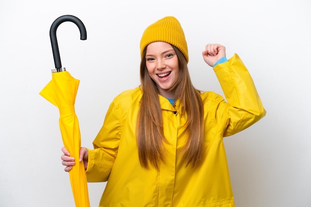 Jovem mulher caucasiana com casaco à prova de chuva e guarda-chuva isolado no fundo branco comemorando uma vitória