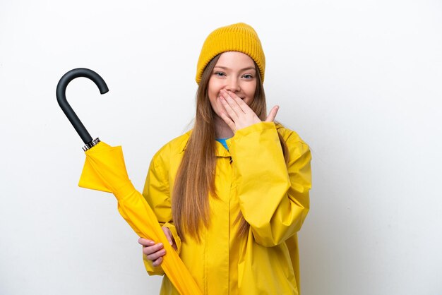 Jovem mulher caucasiana com casaco à prova de chuva e guarda-chuva isolado no fundo branco feliz e sorridente cobrindo a boca com a mão