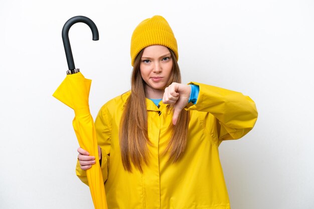 Jovem mulher caucasiana com casaco à prova de chuva e guarda-chuva isolado no fundo branco, mostrando o polegar para baixo com expressão negativa