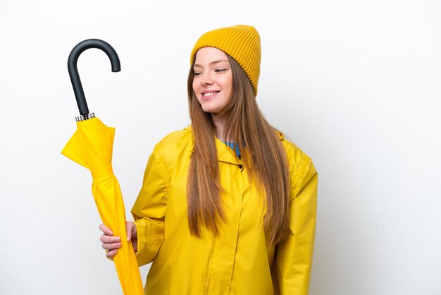 Jovem mulher caucasiana com casaco à prova de chuva e guarda-chuva isolado no fundo branco, olhando para o lado e sorrindo