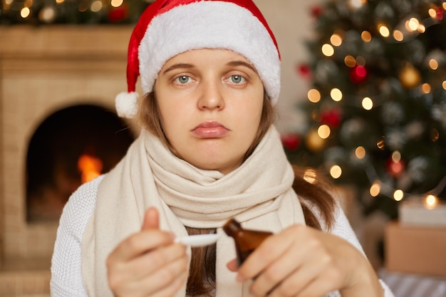 Jovem mulher caucasiana com chapéu de Natal com calda de tosse, doença devido a vírus ou infecção, com expressão chateada, embrulhada em lenço de guerra, posa na sala com enfeites de ano novo