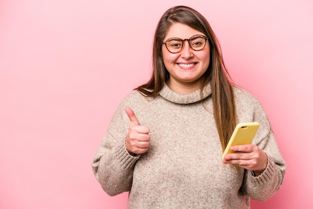 Jovem mulher caucasiana com excesso de peso segurando um telefone celular isolado no fundo rosa sorrindo e levantando o polegar