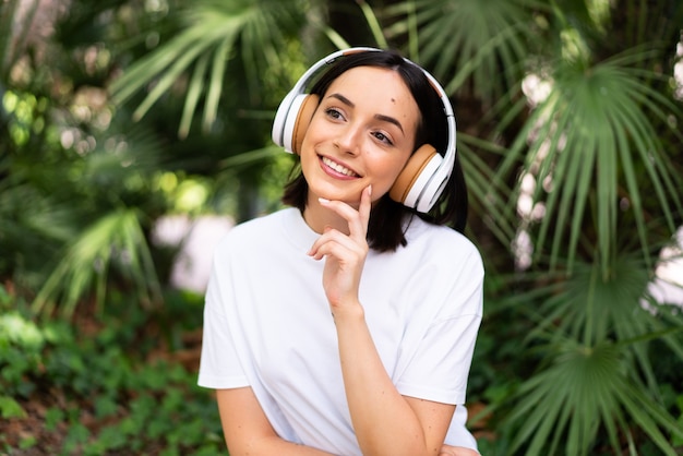 Jovem mulher caucasiana com fones de ouvido ao ar livre, pensando em uma ideia enquanto olha para cima