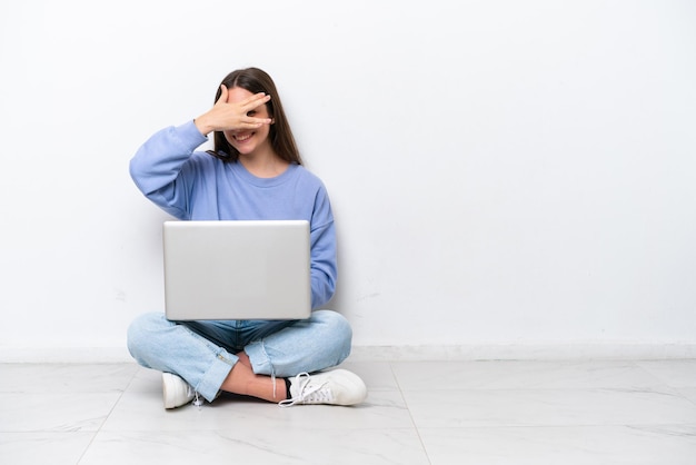 Jovem mulher caucasiana com laptop sentado no chão isolado no fundo branco cobrindo os olhos pelas mãos e sorrindo