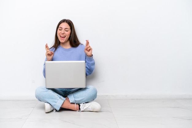 Jovem mulher caucasiana com laptop sentado no chão isolado no fundo branco com os dedos cruzando