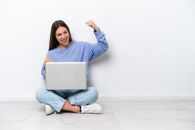 Jovem mulher caucasiana com laptop sentado no chão isolado no fundo branco comemorando uma vitória