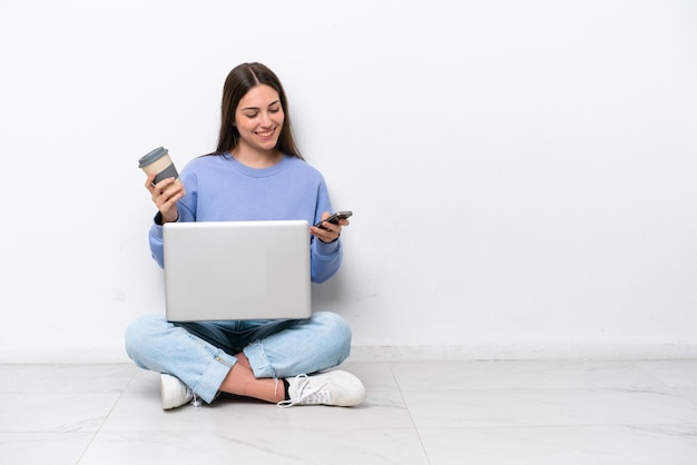 Jovem mulher caucasiana com laptop sentado no chão isolado no fundo branco segurando café para levar e um celular