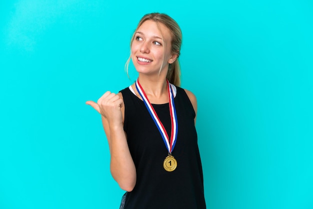 Jovem mulher caucasiana com medalhas isoladas em fundo azul apontando para o lado para apresentar um produto