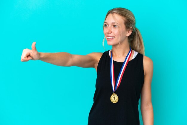 Jovem mulher caucasiana com medalhas isoladas em fundo azul, dando um polegar para cima gesto