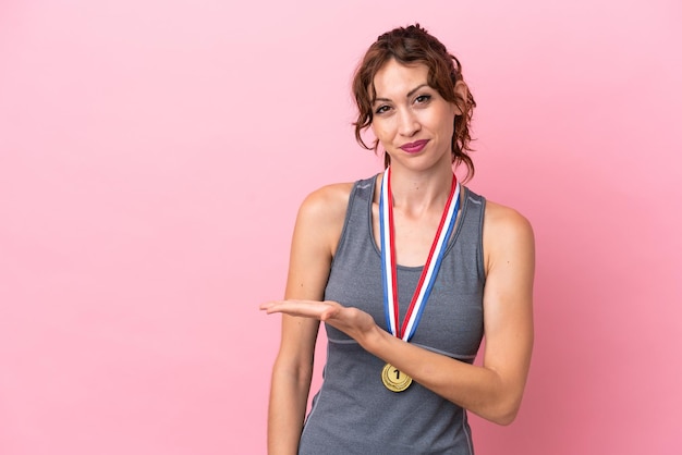 Jovem mulher caucasiana com medalhas isoladas em fundo rosa apresentando uma ideia enquanto olha sorrindo para