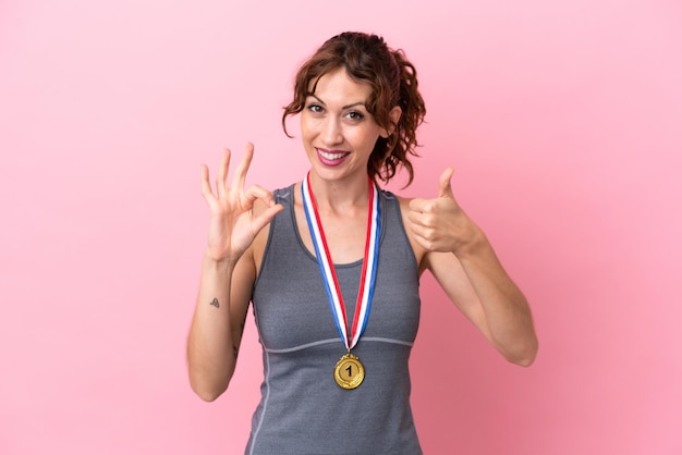 Jovem mulher caucasiana com medalhas isoladas em fundo rosa mostrando sinal de ok e polegar para cima gesto