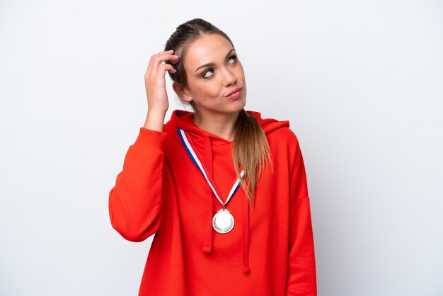 Jovem mulher caucasiana com medalhas isoladas no fundo branco com dúvidas e com expressão de rosto confuso