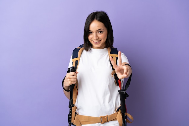 Jovem mulher caucasiana com mochila e bastões de trekking isolados em fundo azul sorrindo e mostrando sinal de vitória