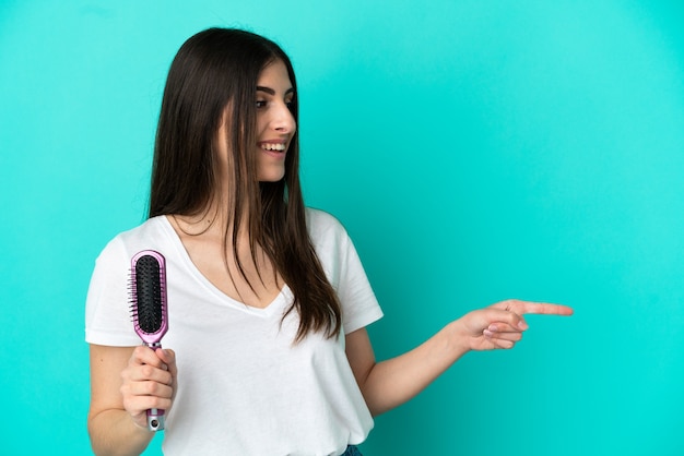 Jovem mulher caucasiana com pente de cabelo isolado em fundo azul apontando para o lado para apresentar um produto