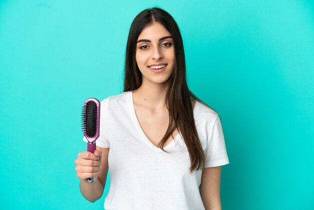 Jovem mulher caucasiana com pente de cabelo isolado em um fundo azul e sorrindo muito