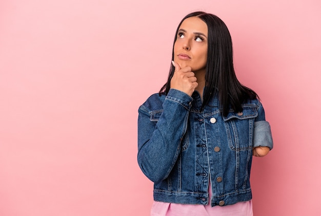 Jovem mulher caucasiana com um braço isolado no fundo rosa, olhando de soslaio com expressão duvidosa e cética.