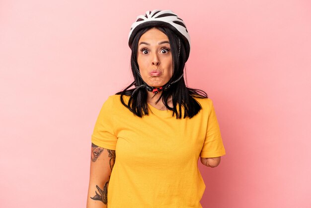 Jovem mulher caucasiana com um braço usando um capacete de bicicleta isolado no fundo rosa encolhe os ombros e abre os olhos confusos.