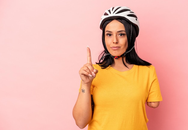 Foto jovem mulher caucasiana com um braço usando um capacete de bicicleta isolado no fundo rosa, mostrando o número um com o dedo.