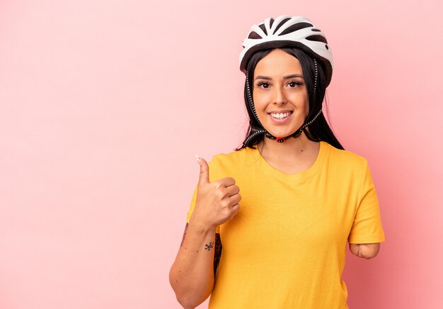 Jovem mulher caucasiana com um braço usando um capacete de bicicleta isolado no fundo rosa, sorrindo e levantando o polegar