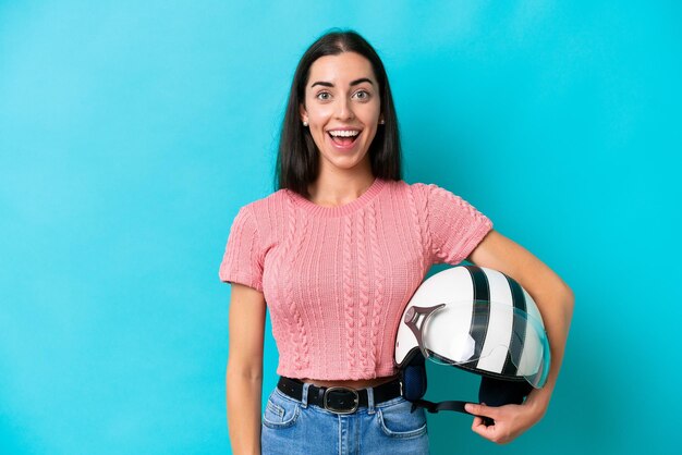 Jovem mulher caucasiana com um capacete de moto isolado em fundo azul com expressão facial de surpresa