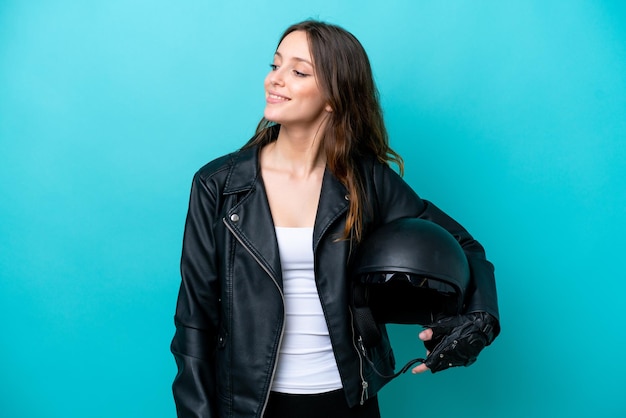 Jovem mulher caucasiana com um capacete de moto isolado em fundo azul, olhando para o lado e sorrindo