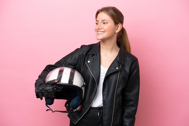 Jovem mulher caucasiana com um capacete de moto isolado em fundo rosa, olhando para o lado e sorrindo