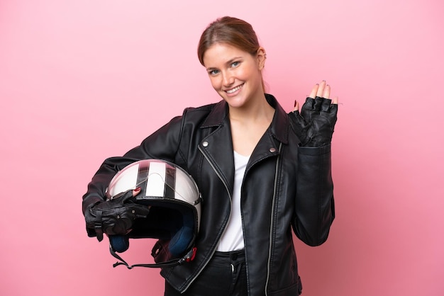 Jovem mulher caucasiana com um capacete de moto isolado em fundo rosa saudando com a mão com expressão feliz