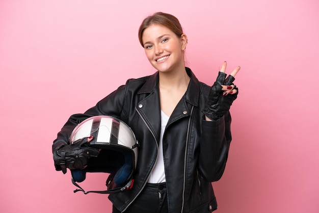 Jovem mulher caucasiana com um capacete de moto isolado em fundo rosa sorrindo e mostrando sinal de vitória