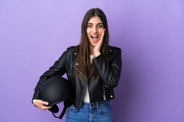 Jovem mulher caucasiana com um capacete de motociclista isolada em um fundo roxo com expressão facial surpresa e chocada