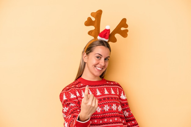 Foto jovem mulher caucasiana com um chapéu de rena de natal, isolado em um fundo amarelo, apontando com o dedo para você, como se fosse um convite para se aproximar.