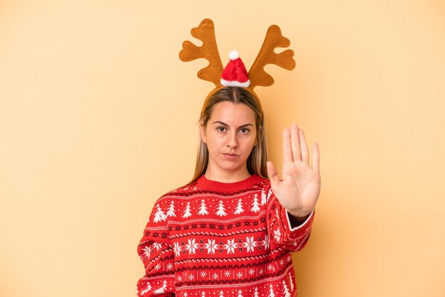 Jovem mulher caucasiana, com um chapéu de rena de Natal, isolado em um fundo amarelo, de pé com a mão estendida, mostrando o sinal de pare, impedindo-o.