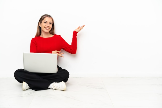 Jovem mulher caucasiana com um laptop sentada no chão, estendendo as mãos para o lado para convidar para vir