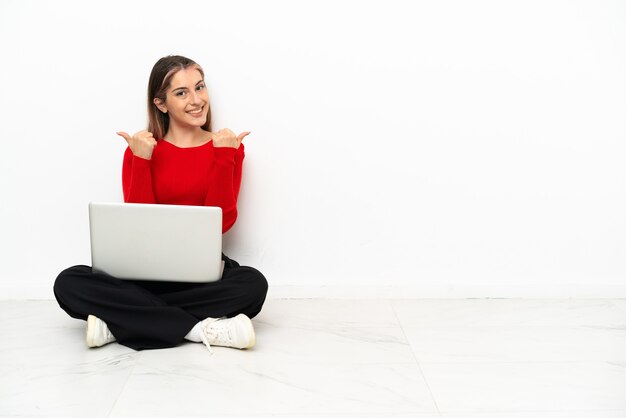 Jovem mulher caucasiana com um laptop sentada no chão, fazendo gestos de polegar para cima e sorrindo